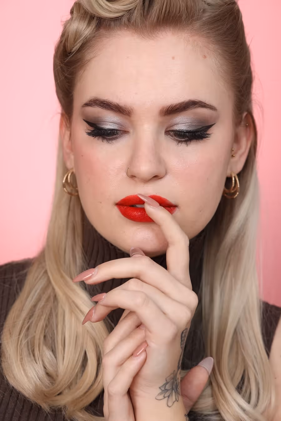 Frau mit roten Lippen, silbernem Lidschatten und langen blonden Haaren vor rosa Hintergrund, mit Tattoo auf der Hand.