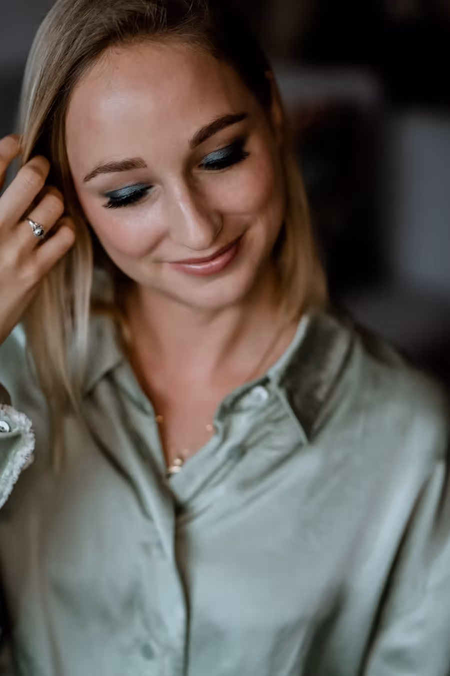 Junge Frau mit blondem Haar und grünem Hemd, die lächelnd nach unten schaut und sich an den Haaren berührt.