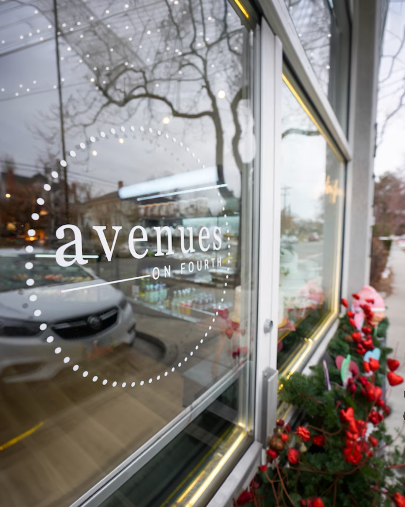 Glass door with white circular logo reading 'avenues on fourth' with reflections of street and parked car, and holiday decorations with red berries along the sidewalk.