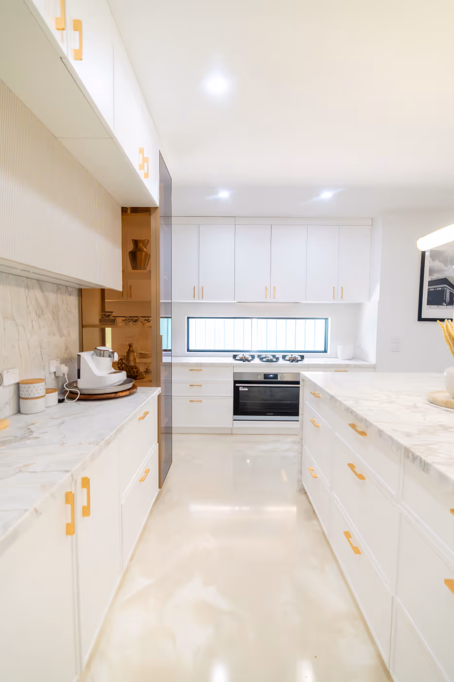 Bright modern kitchen with white cabinets, gold handles, marble countertops, and a built-in stove below a wide window.