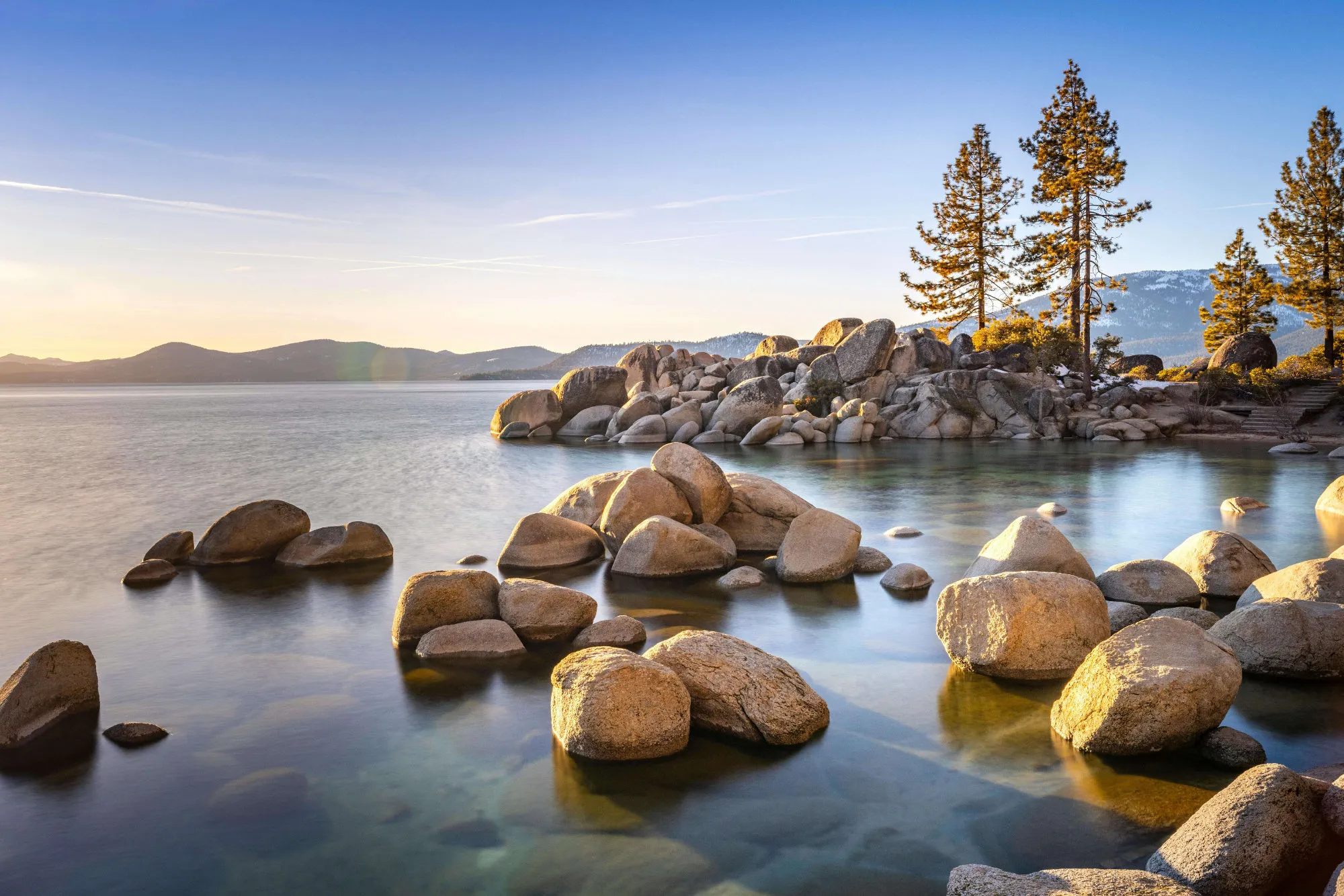 Lake with clear water, large boulders, pine trees, and mountains under a blue sky at sunset.