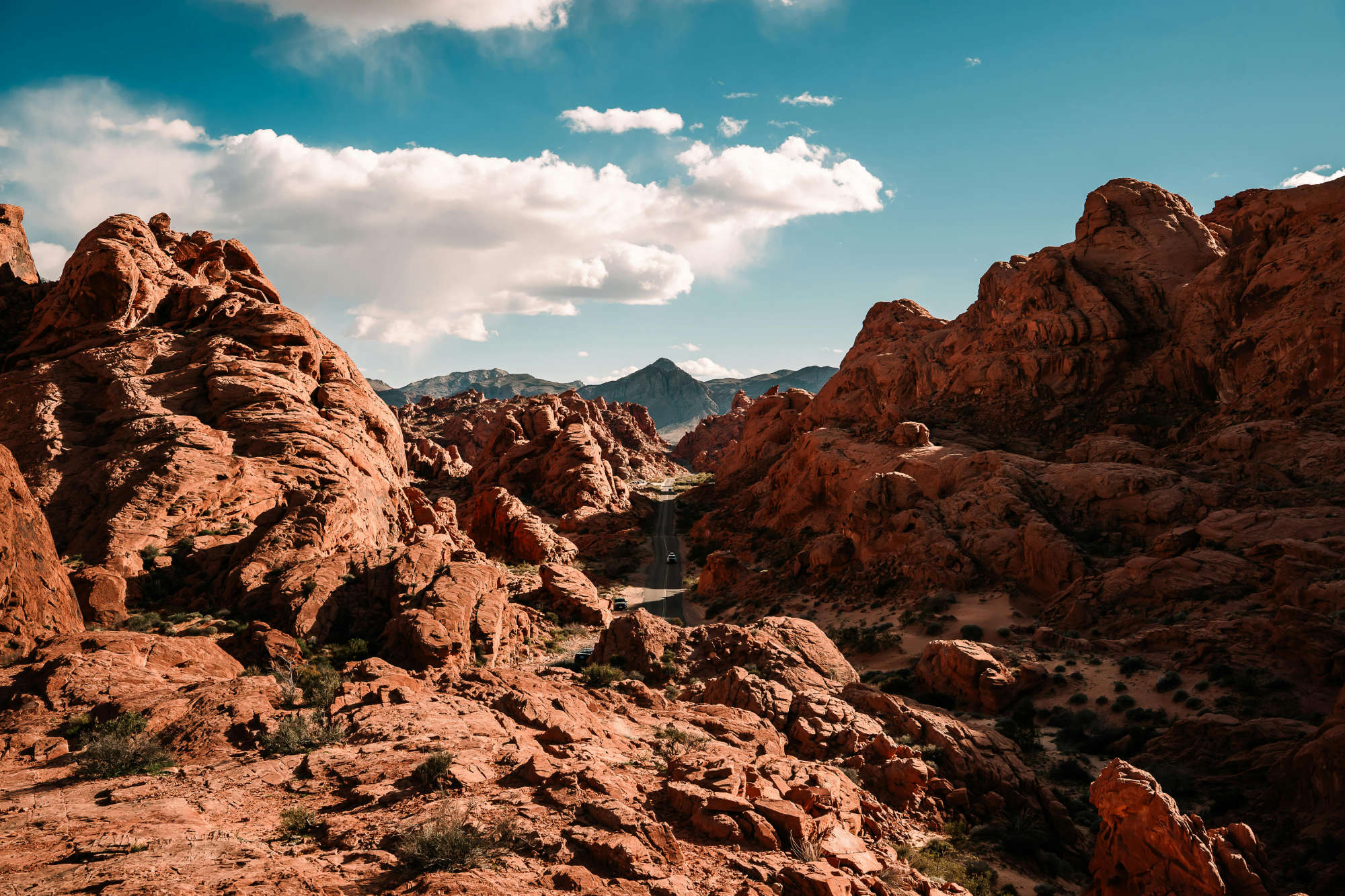 Desert road cutting through red rocky formations under a partly cloudy blue sky.