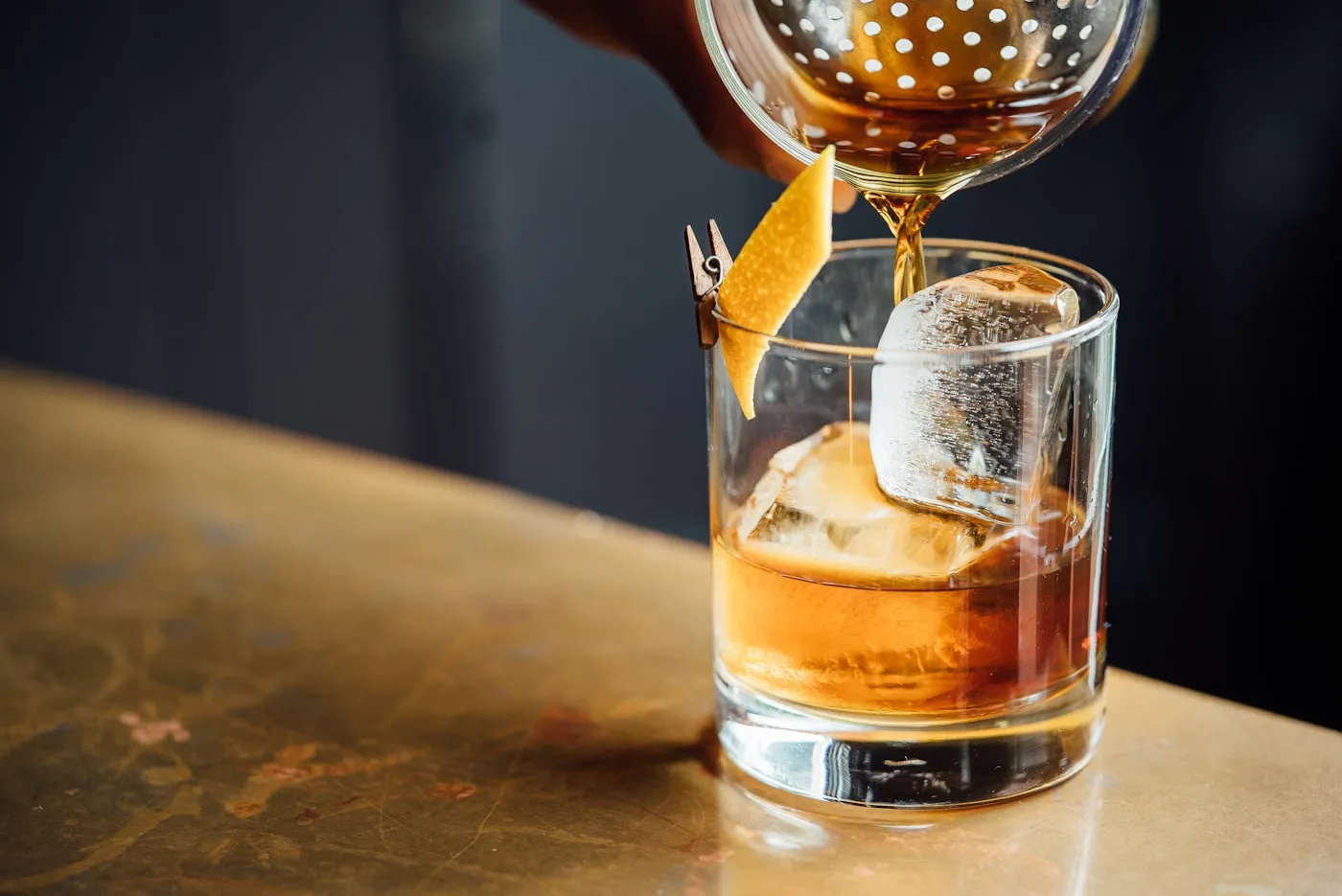 Amber cocktail with ice cubes and an orange peel garnish being poured from a strainer.