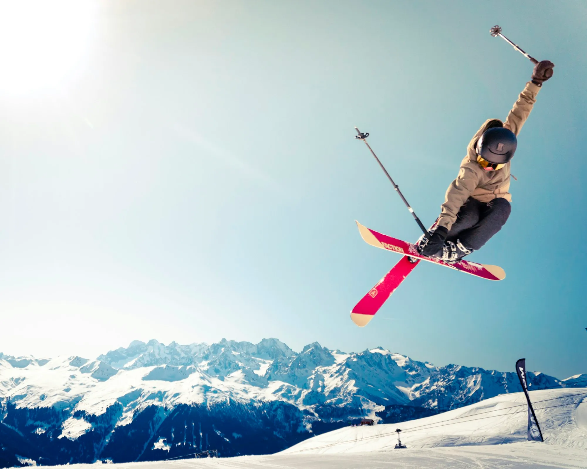 Skier in mid-air performing a jump over snowy slopes with mountains in the background under blue sky.