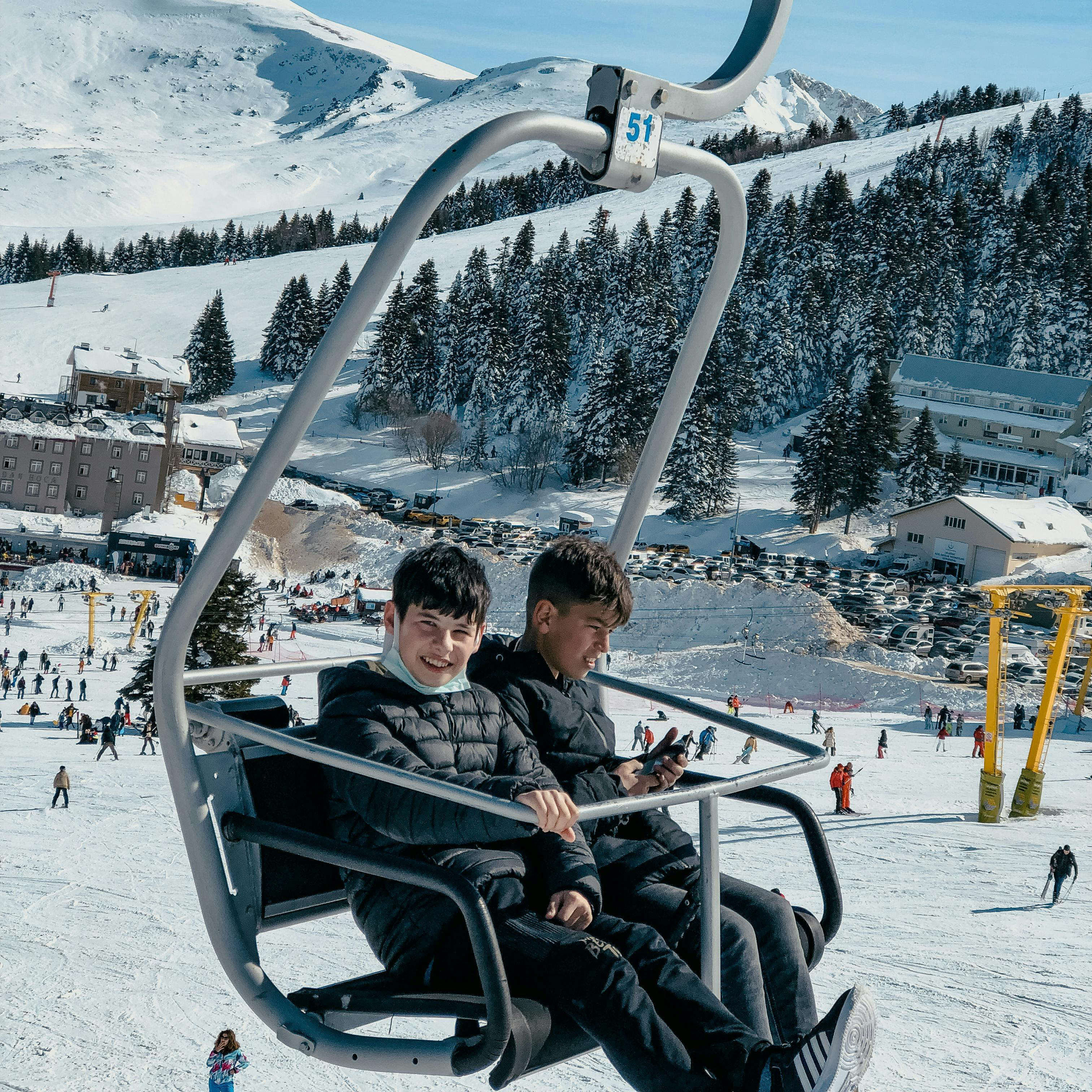 Two boys in black jackets riding a ski lift over snowy slopes and pine trees with mountains in the background.