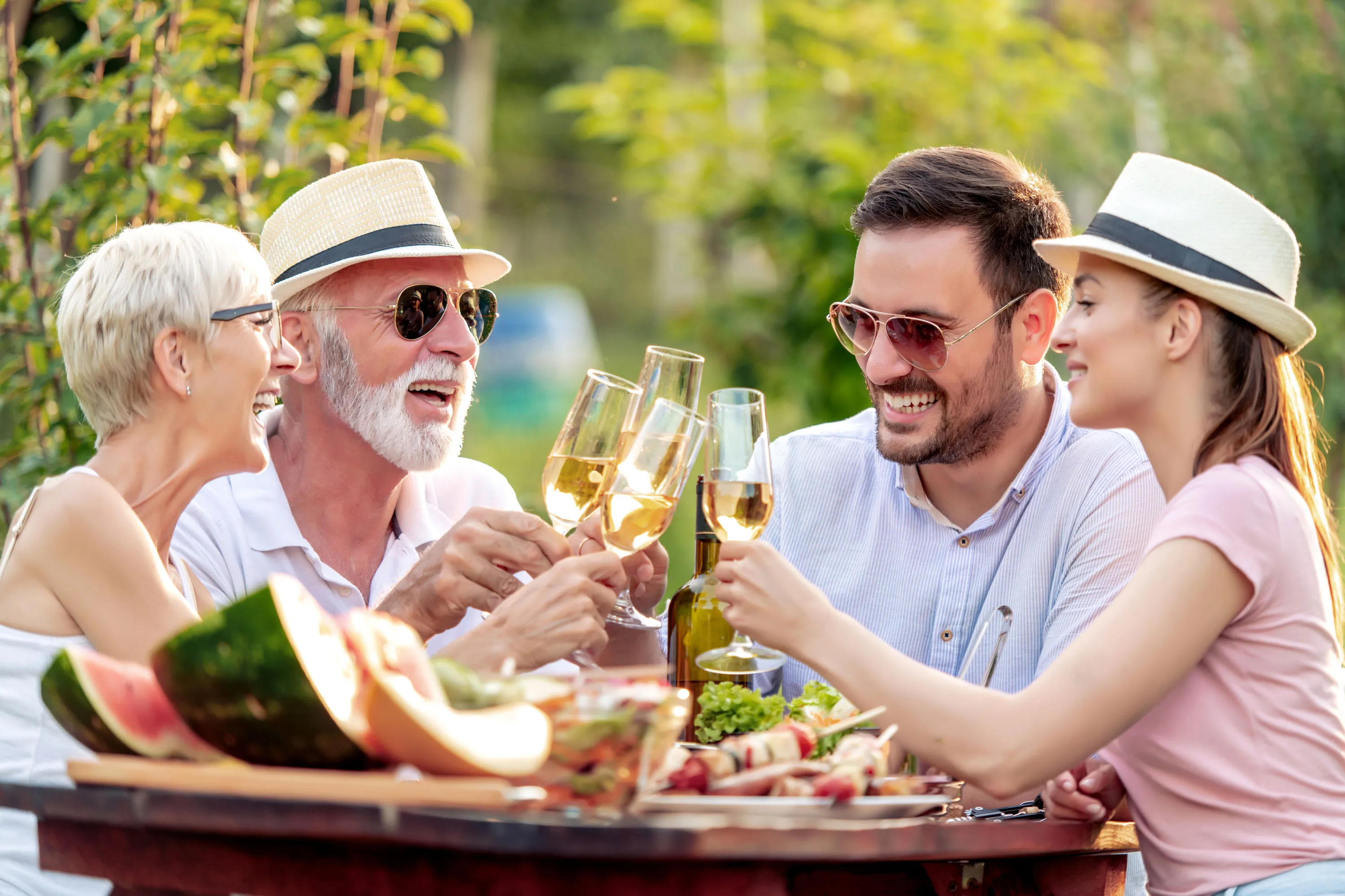 Four people outdoors toasting with wine glasses around a table with food and watermelon slices.