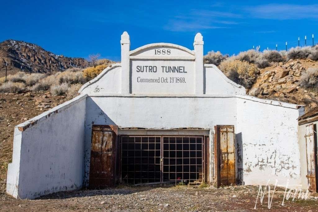 Entrance of the Sutro Tunnel with white facade and rusted metal gates against a mountainous background.