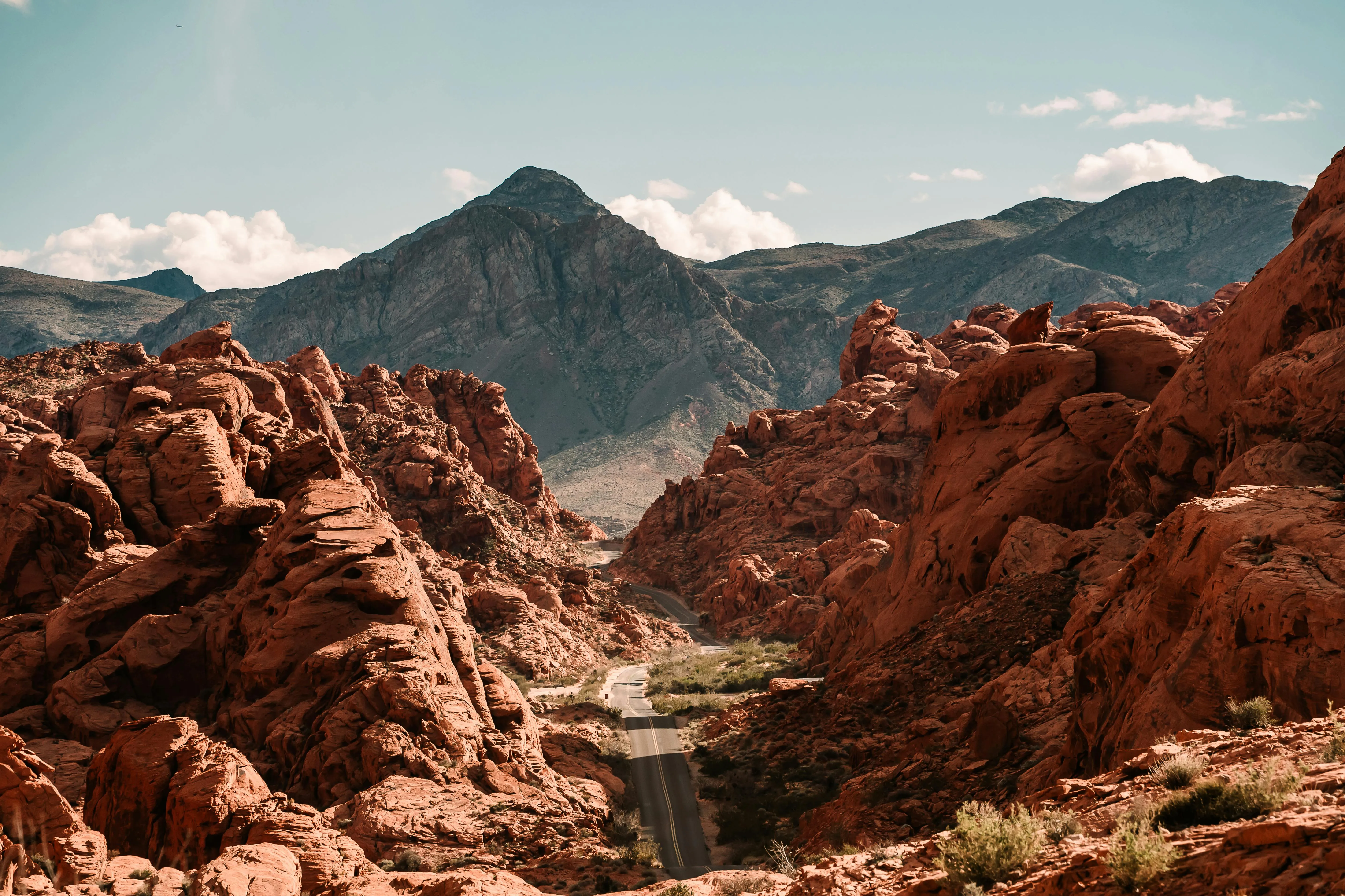 Road winding through reddish rock formations with a mountain range and partly cloudy sky in the background.