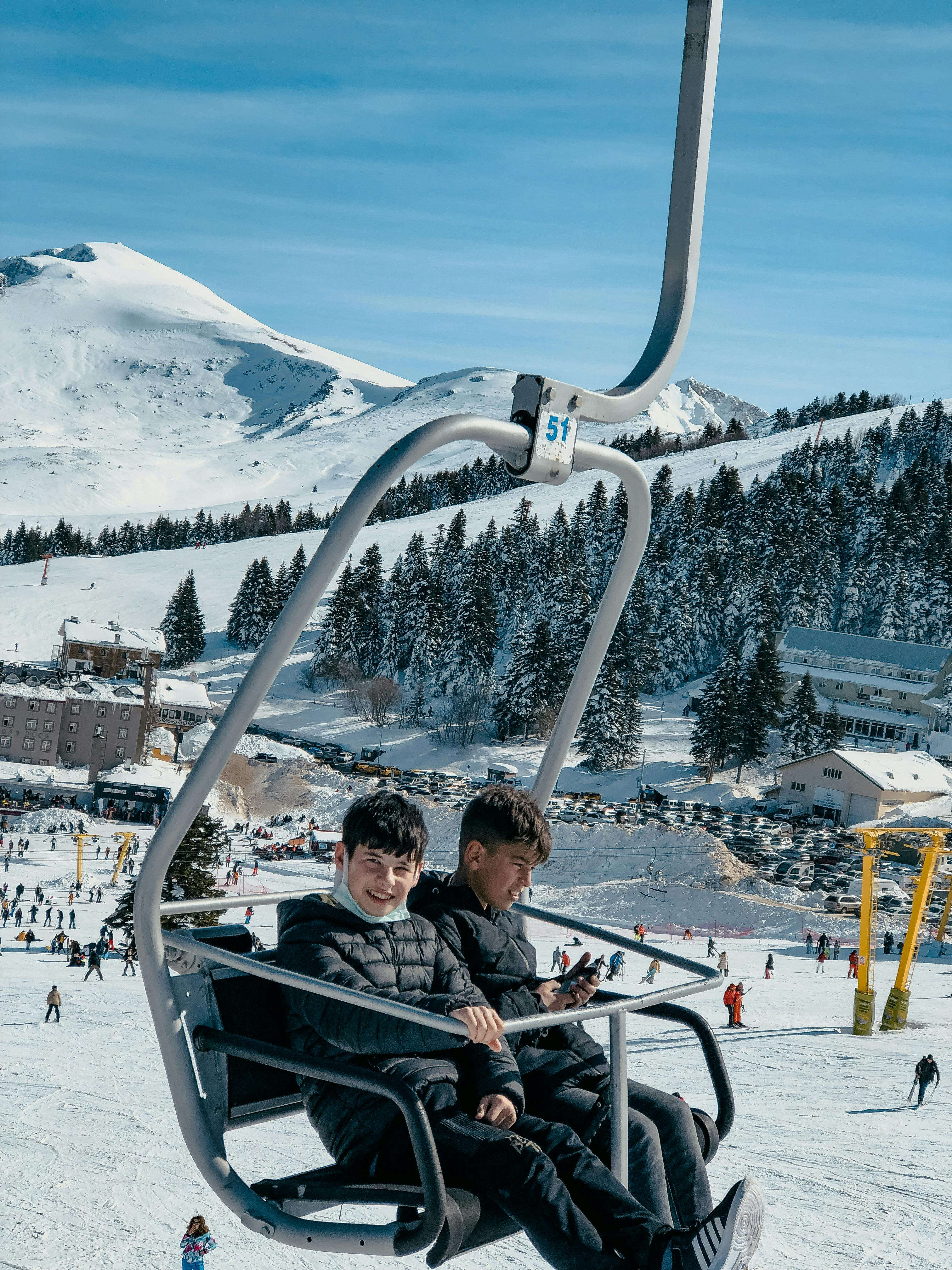 Two boys in black jackets sitting on ski lift over snowy ski slope with trees and mountains.