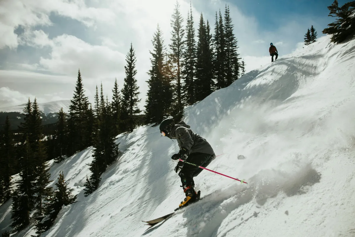 Skier in black gear skiing down a snowy slope with pine trees and a person standing above.
