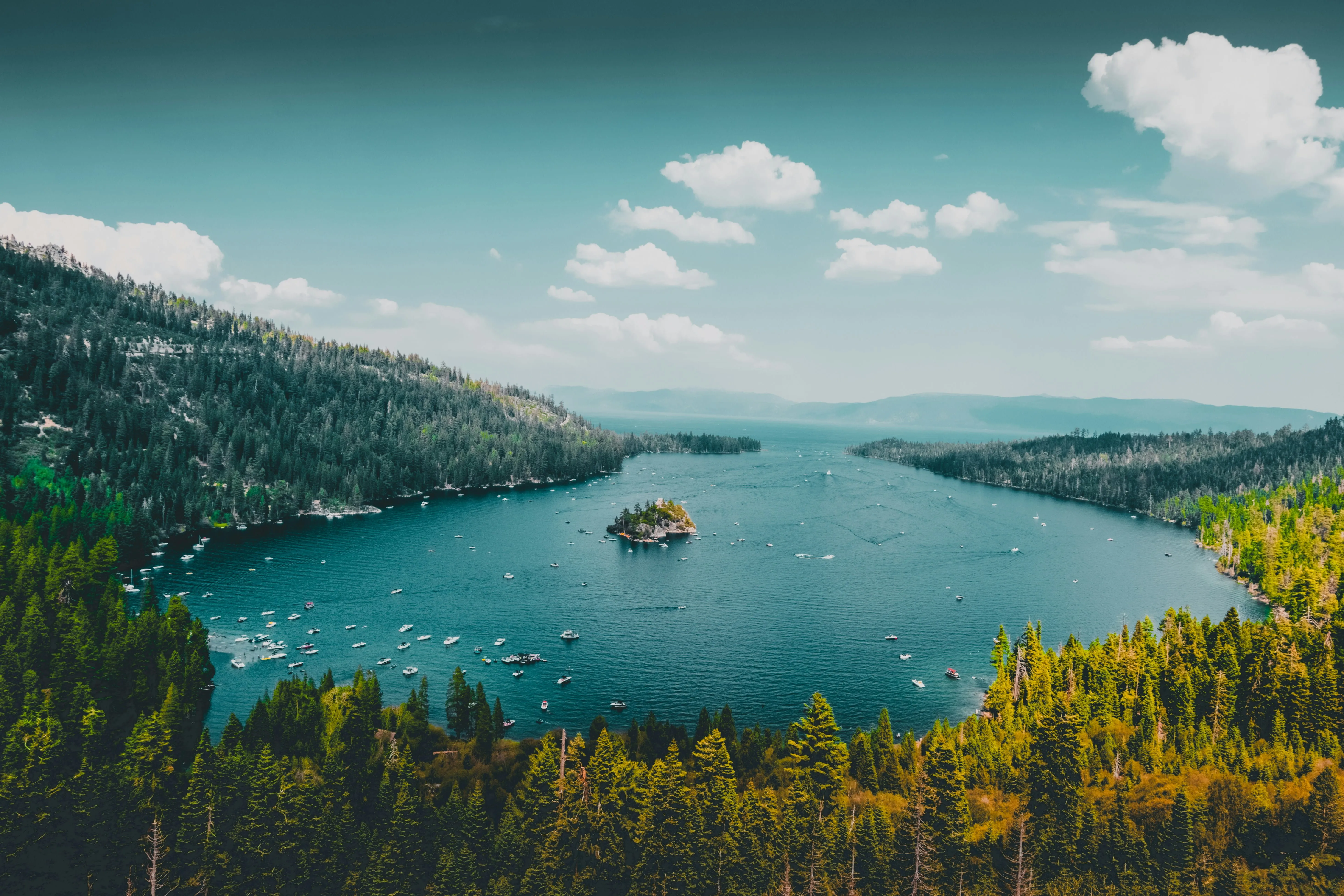 Lake surrounded by forested hills with boats on the water under a partly cloudy sky.
