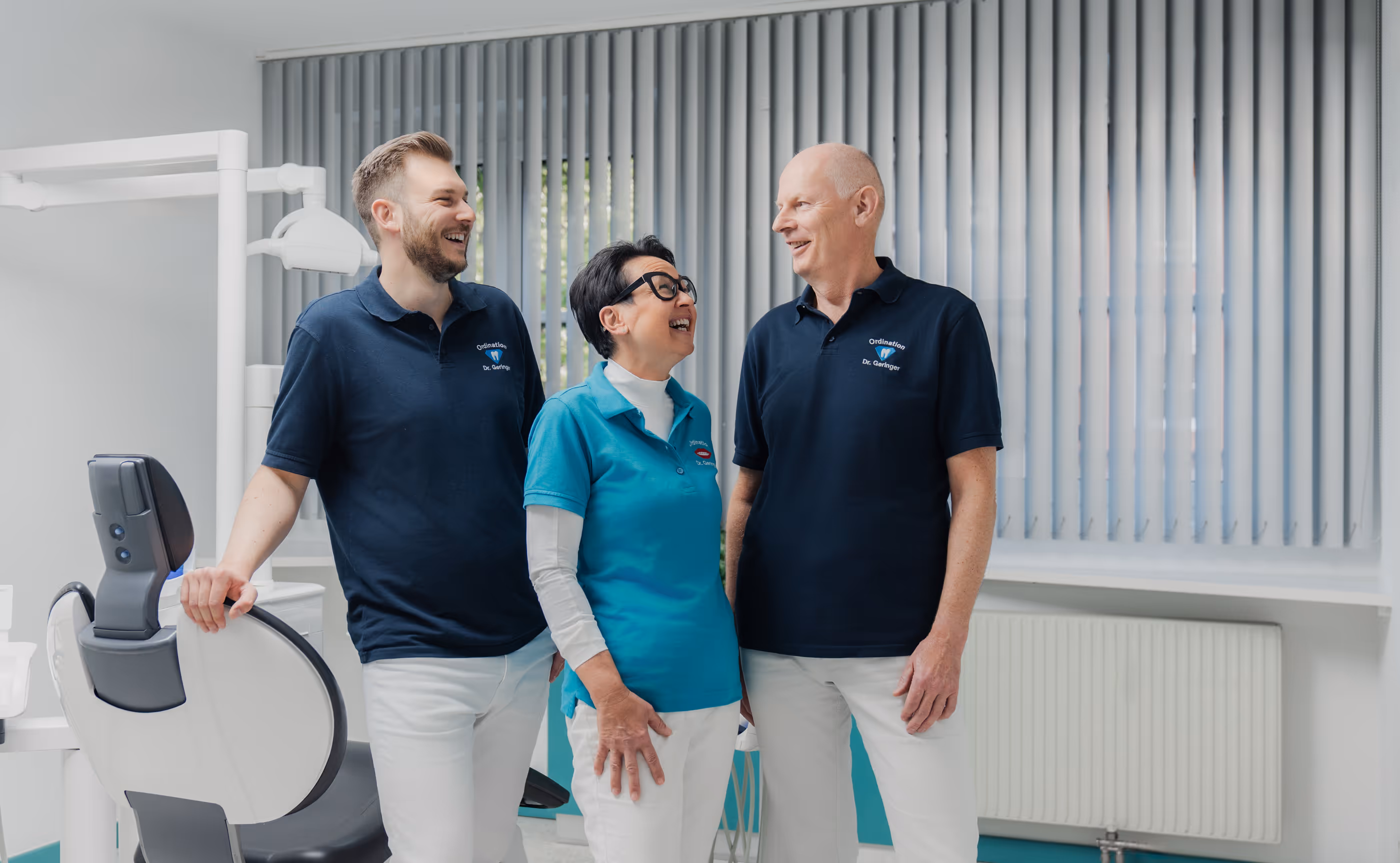 Drei lächelnde Zahnärzte in blauen Poloshirts stehen zusammen in einer modernen Praxis.