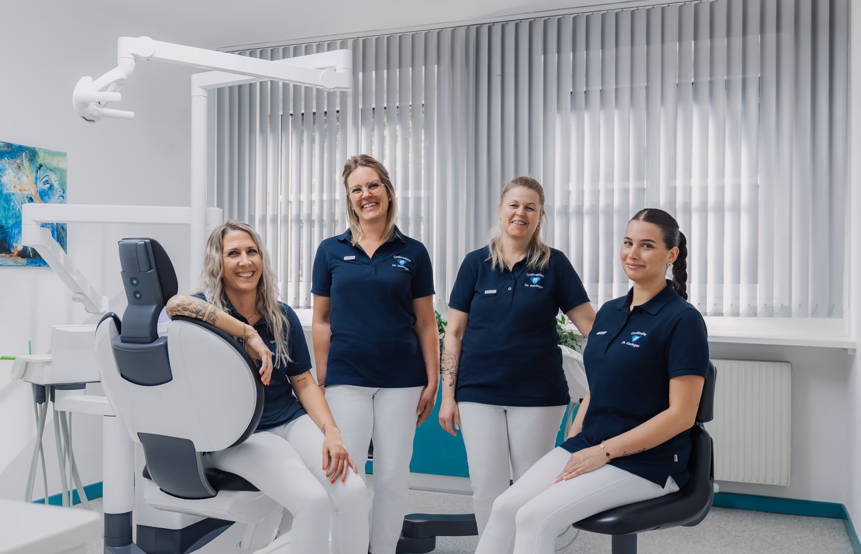 Vier Frauen in blauen Poloshirts und weißen Hosen in einer Zahnarztpraxis, lächelnd.