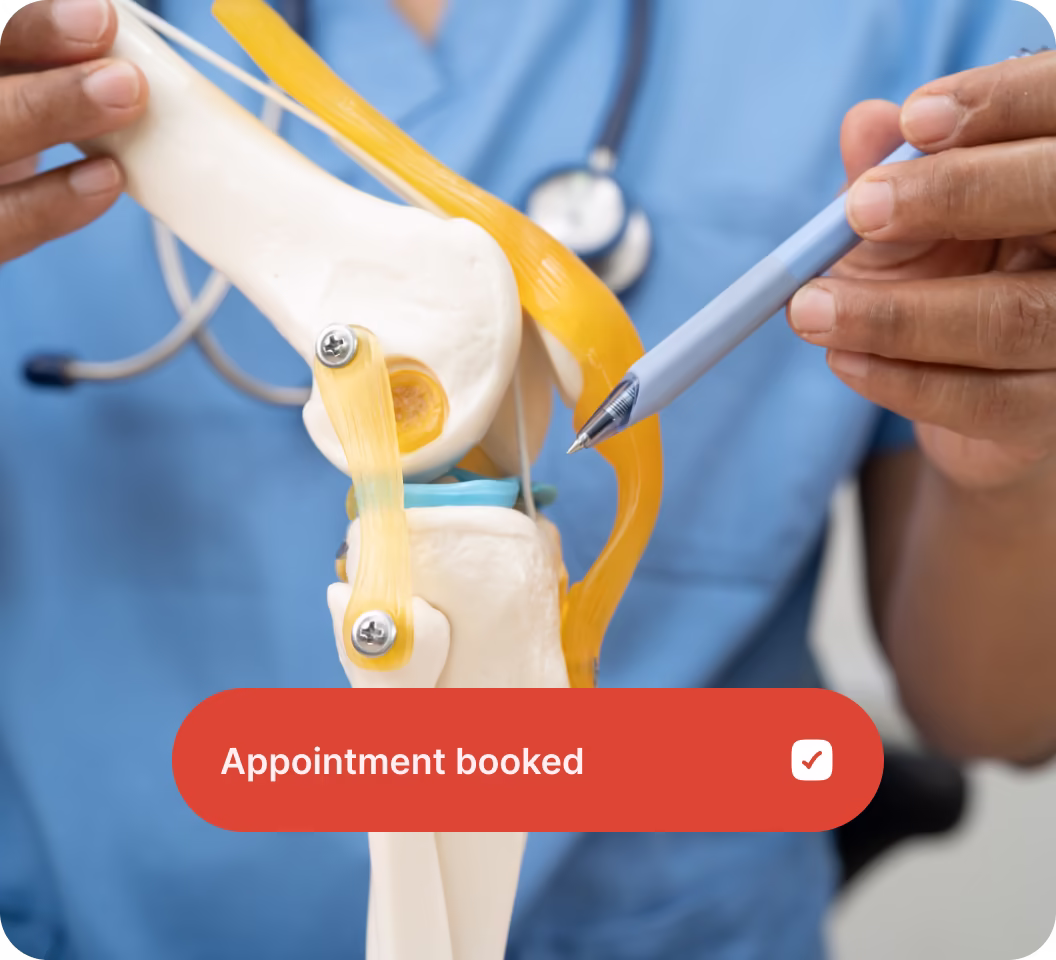 Medical professional in blue scrubs pointing at a knee joint model with a pen, with a stethoscope in the background and an appointment booked notification overlay.