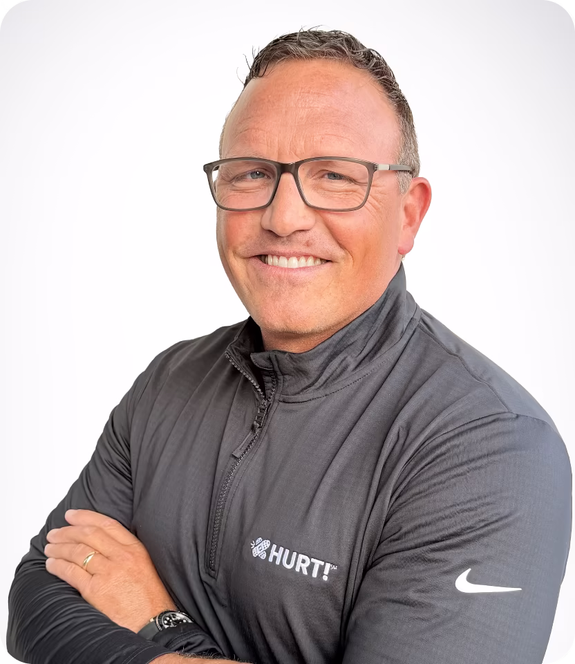 Smiling man wearing glasses and a black Nike zip-up shirt with HURT! logo, arms crossed.