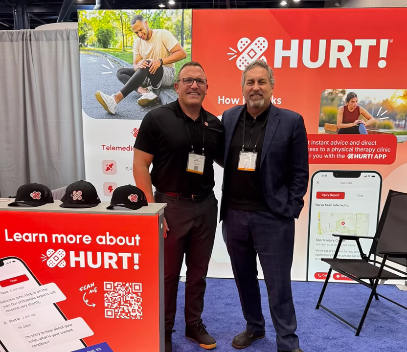 Two men smiling and standing in front of a red and white HURT! app promotional booth with hats on a table and informational graphics about telemedicine and physical therapy.