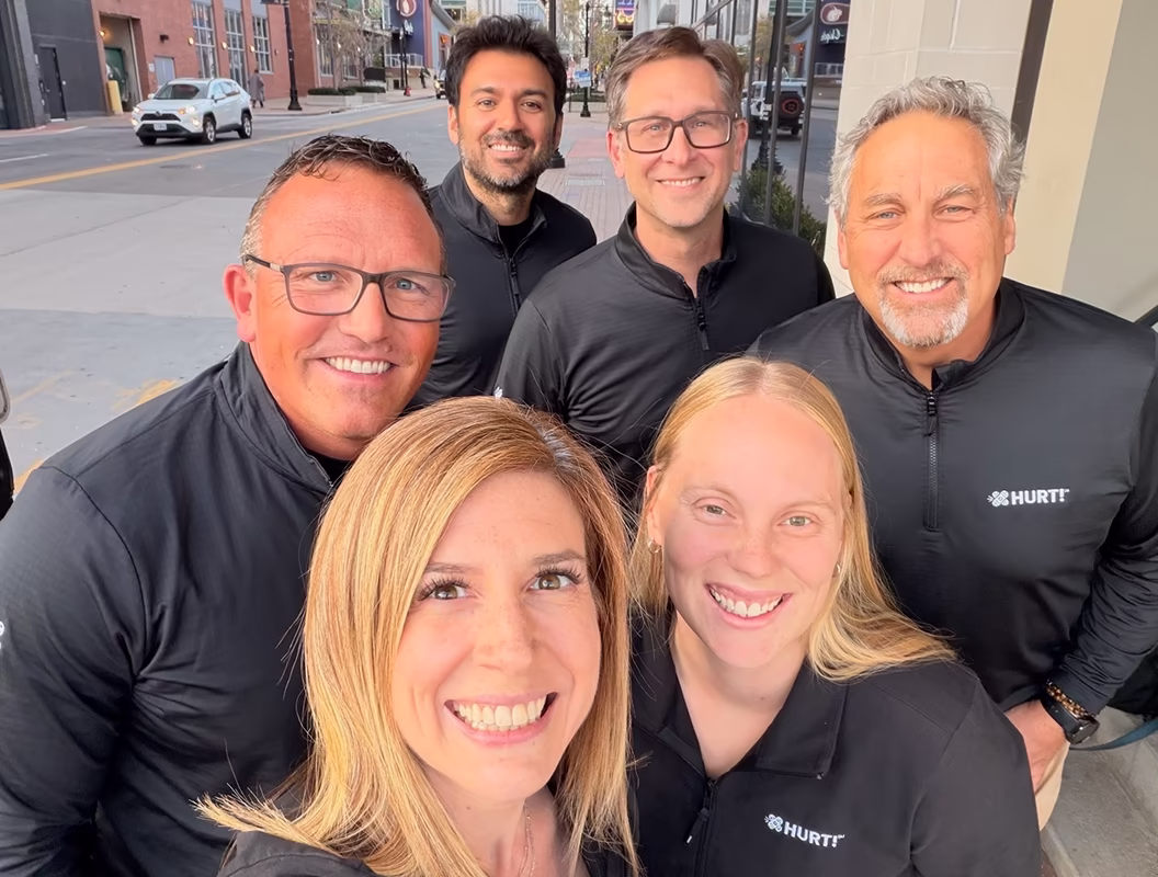Group of six smiling people wearing black zip-up shirts with HURT! logo standing outdoors on a city street.