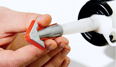 Hand holding a caulking gun nozzle attachment (REGGA) with orange-grey tip while applying silicone