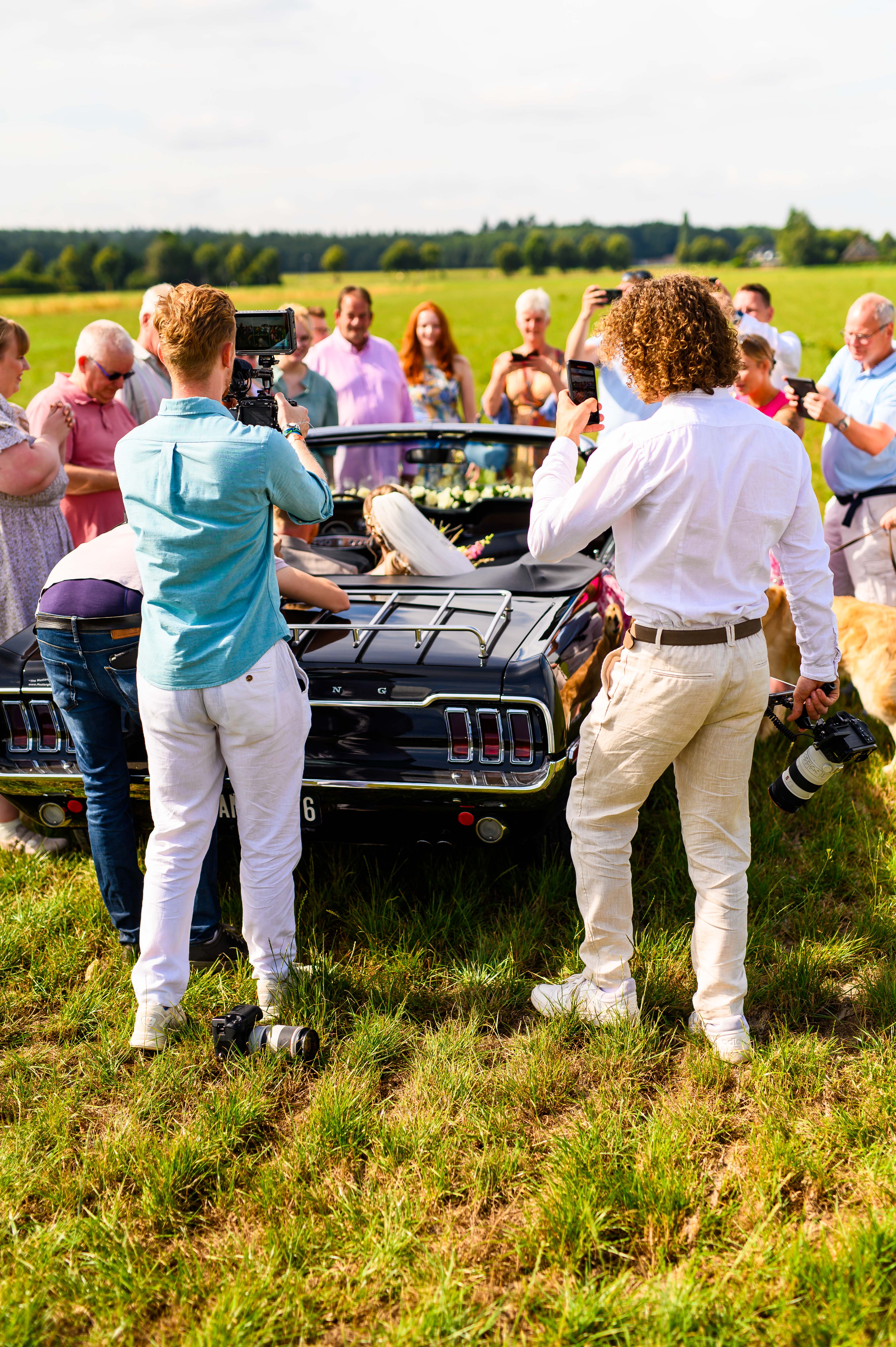 Groep mensen fotografeert bruid in een zwarte cabriolet op een groen veld op een zonnige dag.