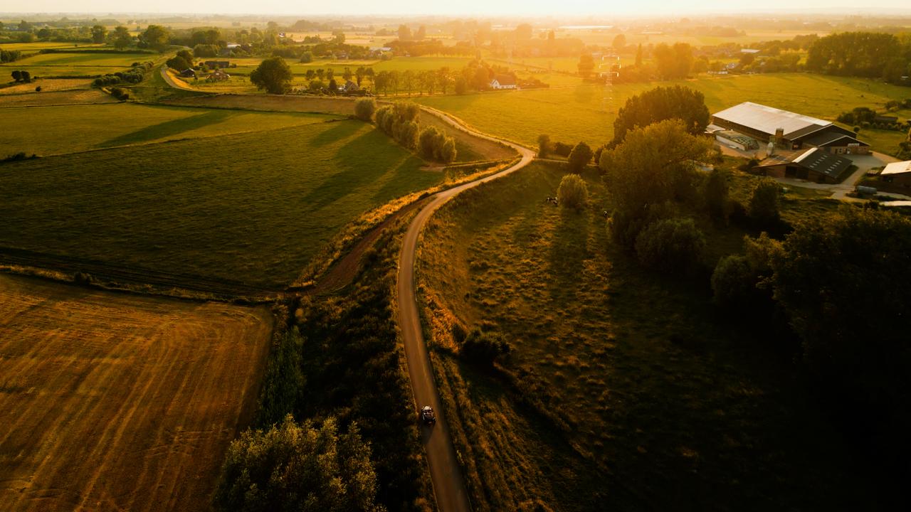 Landschap met weilanden, boerderij en een kronkelende weg in warm zonlicht bij ondergang.