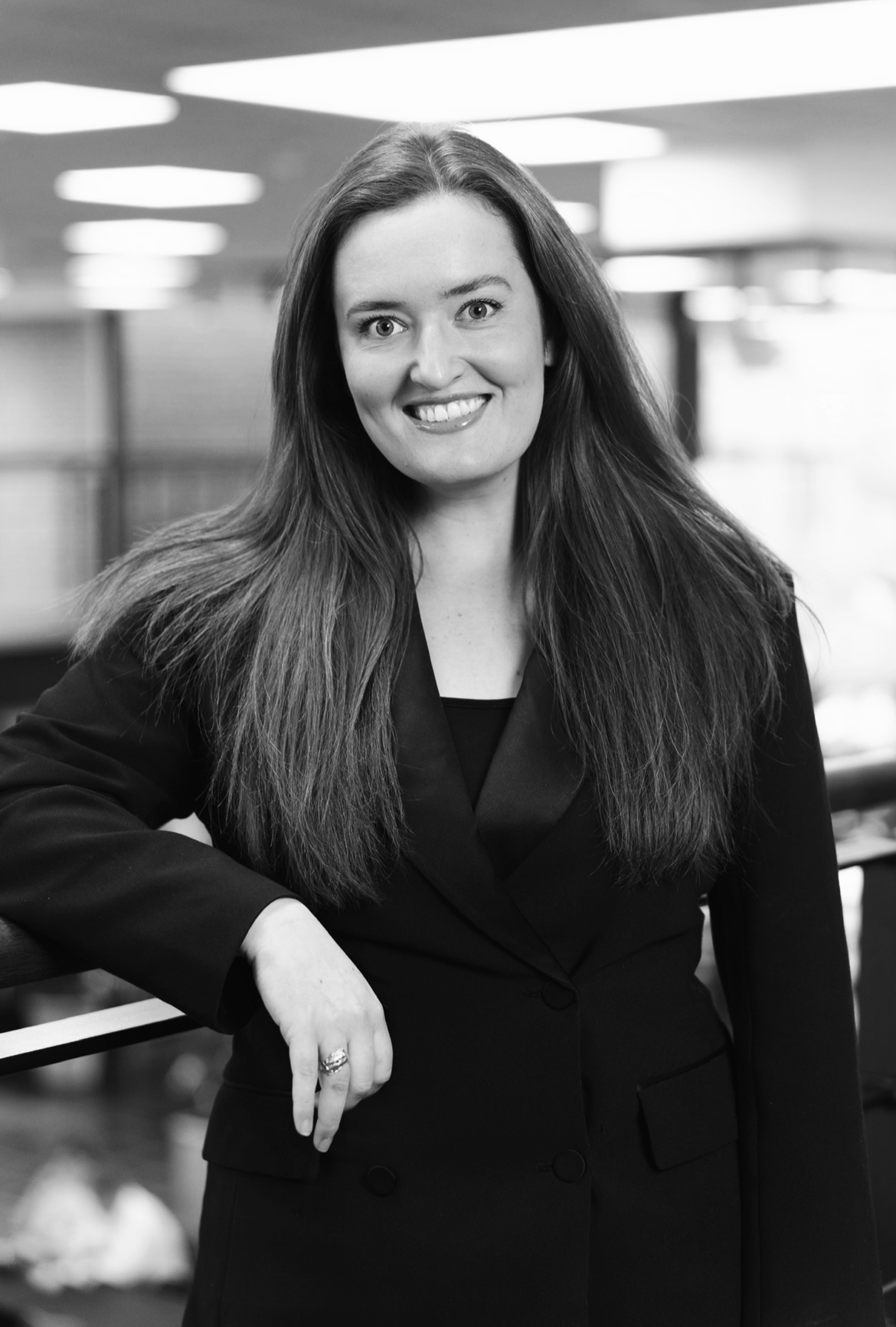 Sort-hvid portræt af en smilende kvinde med langt hår iført en blazer og stående med armen hvilende på en gelænder.