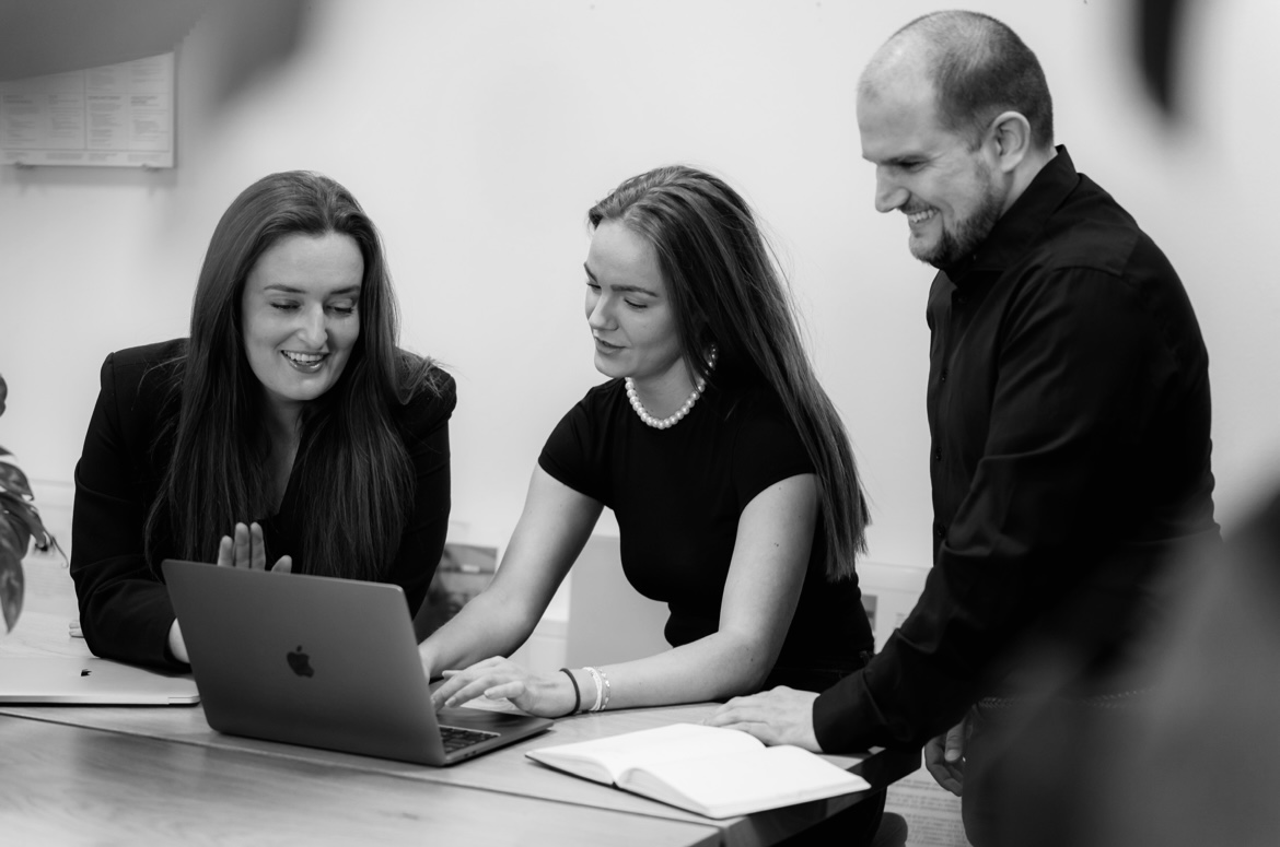 Tre personer, to kvinder og en mand, arbejder sammen og diskuterer noget på en bærbar computer ved et bord.