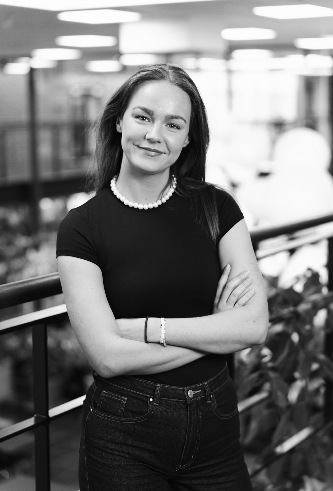Sort-hvidt portræt af en ung kvinde med perlehalskæde og mørkt tøj, stående med armene krydset og smilende.