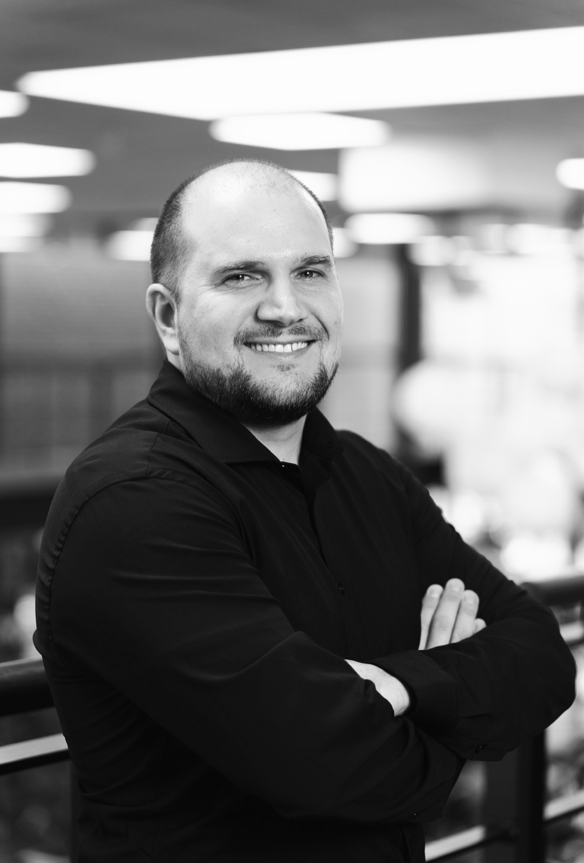 Smilende mand med kort hår og skæg iført sort skjorte, poserer med armene over kors i et kontormiljø.