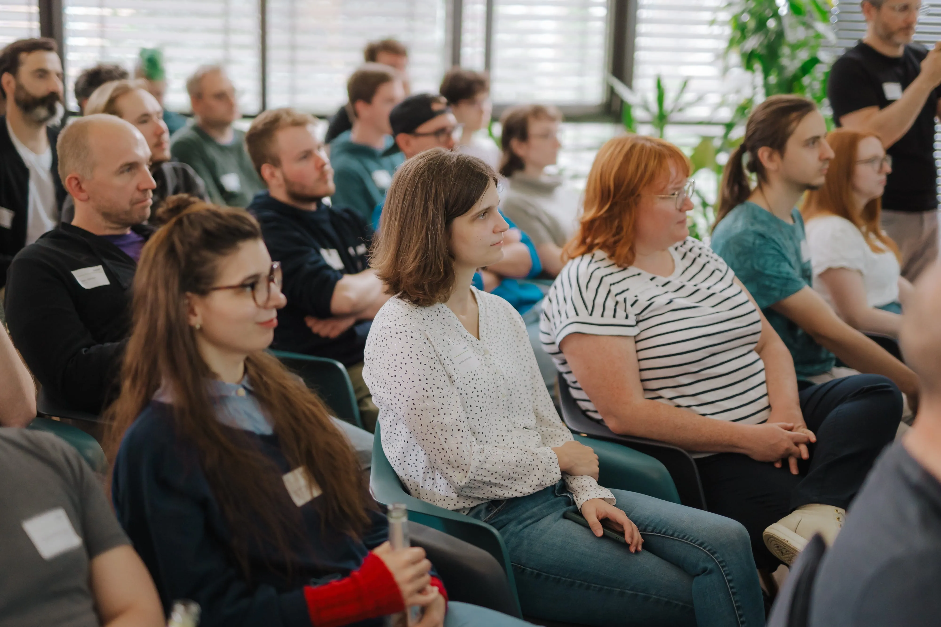 Gruppe von Menschen sitzt aufmerksam bei einem Vortrag in einem hellen Raum mit Pflanzen.