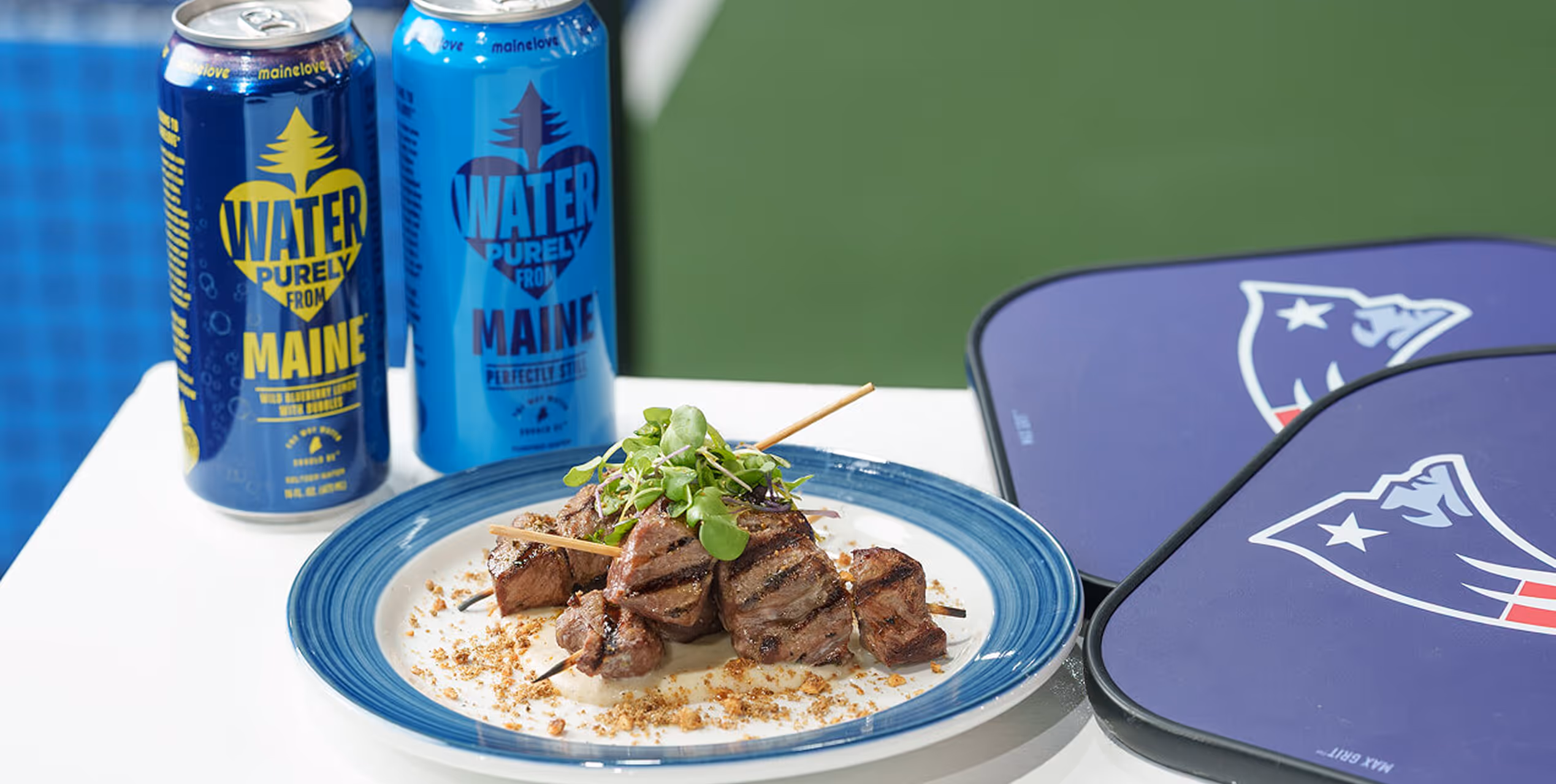 Grilled meat skewers garnished with greens on a blue-rimmed plate with two blue cans of Water from Maine and two purple Patriots folding chairs in the background.