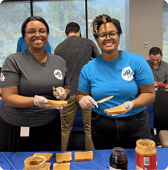 Two smiling women wearing gloves spreading peanut butter on bread slices at a table with peanut butter and jelly jars.