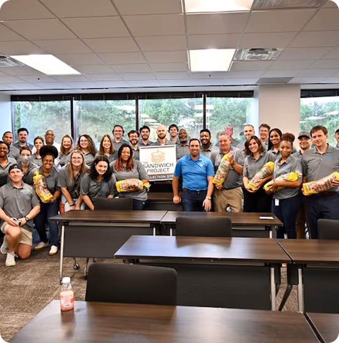 Group of people in gray shirts holding large wrapped sandwiches in a meeting room.