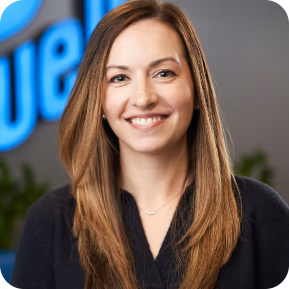 Smiling woman with long brown hair wearing a black top in an office setting with blurred background.