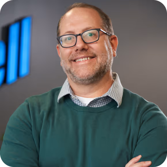 Smiling man with glasses and beard wearing green sweater and checkered shirt in an office setting.