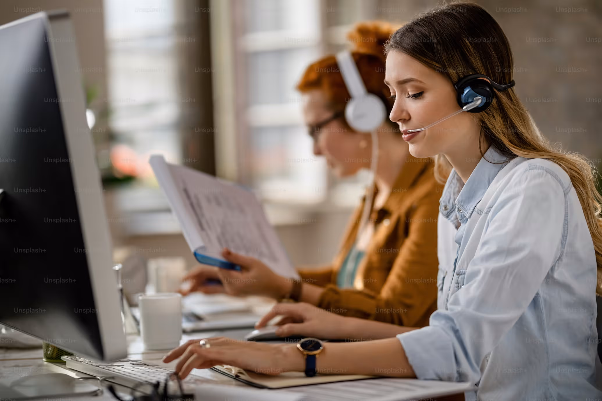 Two women wearing headsets working at computers in an office, one typing, the other reading documents.
