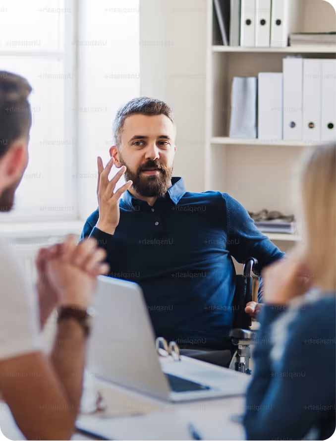 Man in wheelchair gesturing and talking to two seated colleagues at a table with laptop.