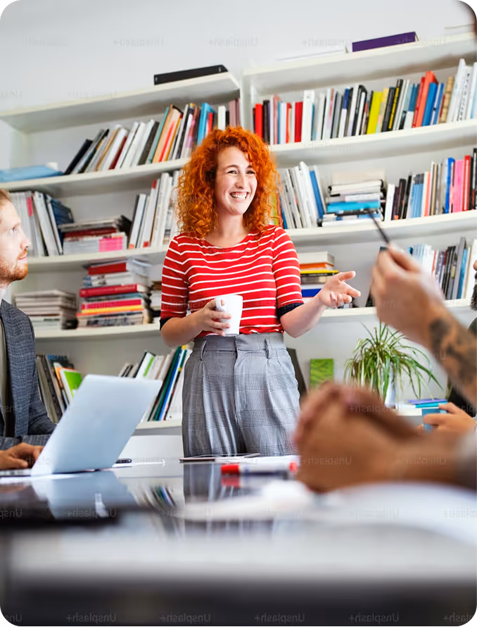 Woman with curly red hair in red striped shirt speaking and holding a cup in a meeting room with bookshelves.