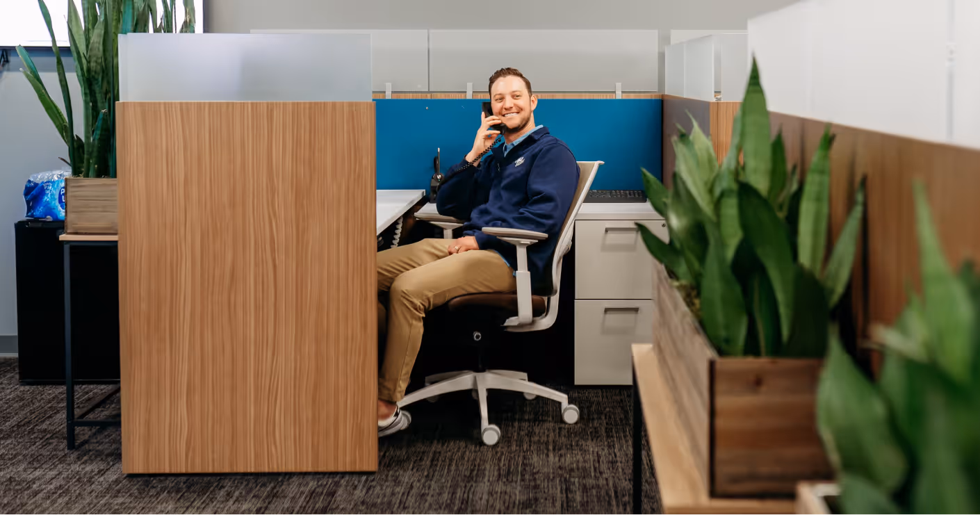 Man sitting in office cubicle smiling while talking on a phone, with plants nearby.