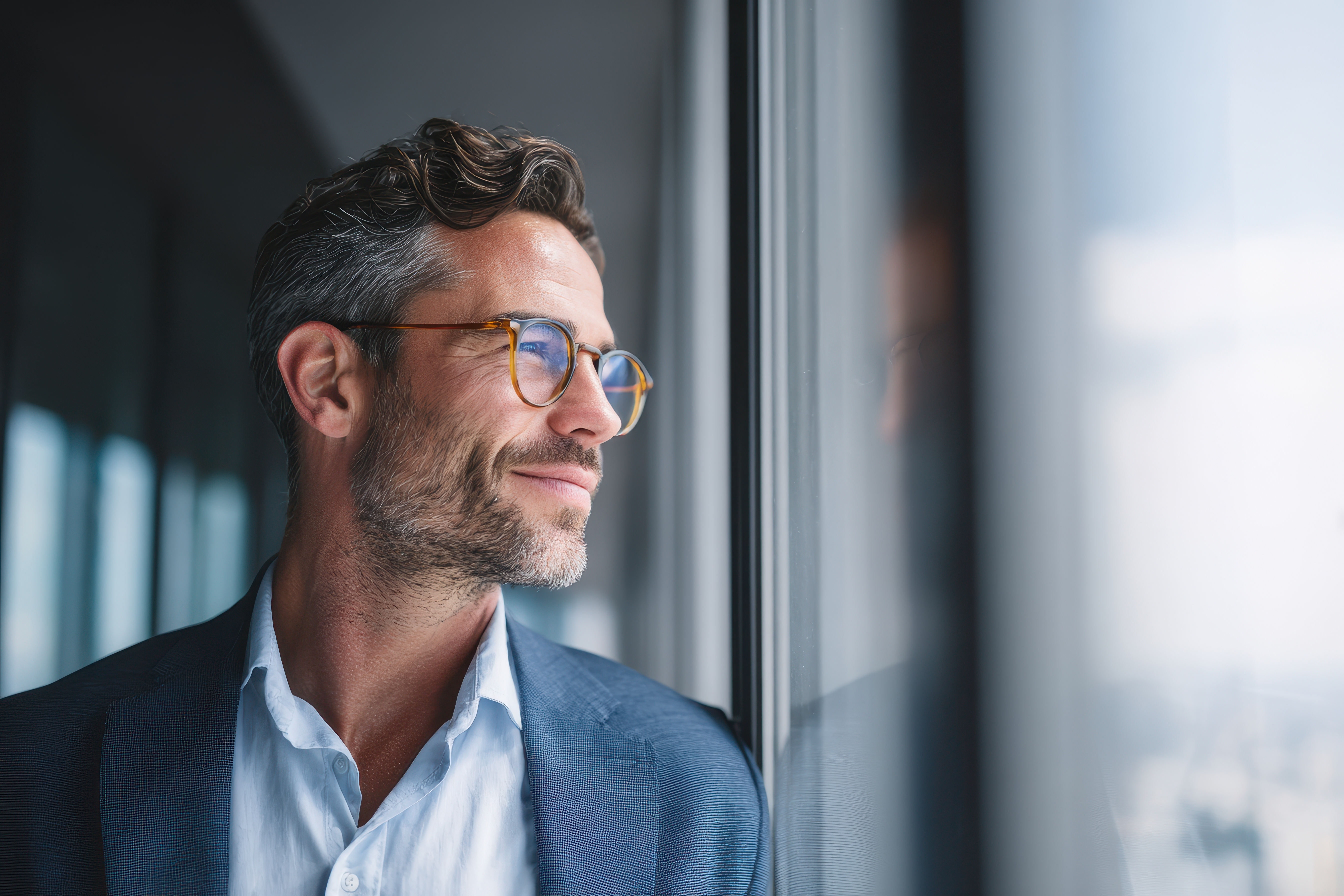 Smiling man with glasses wearing a blue blazer looking out a window with soft natural light.
