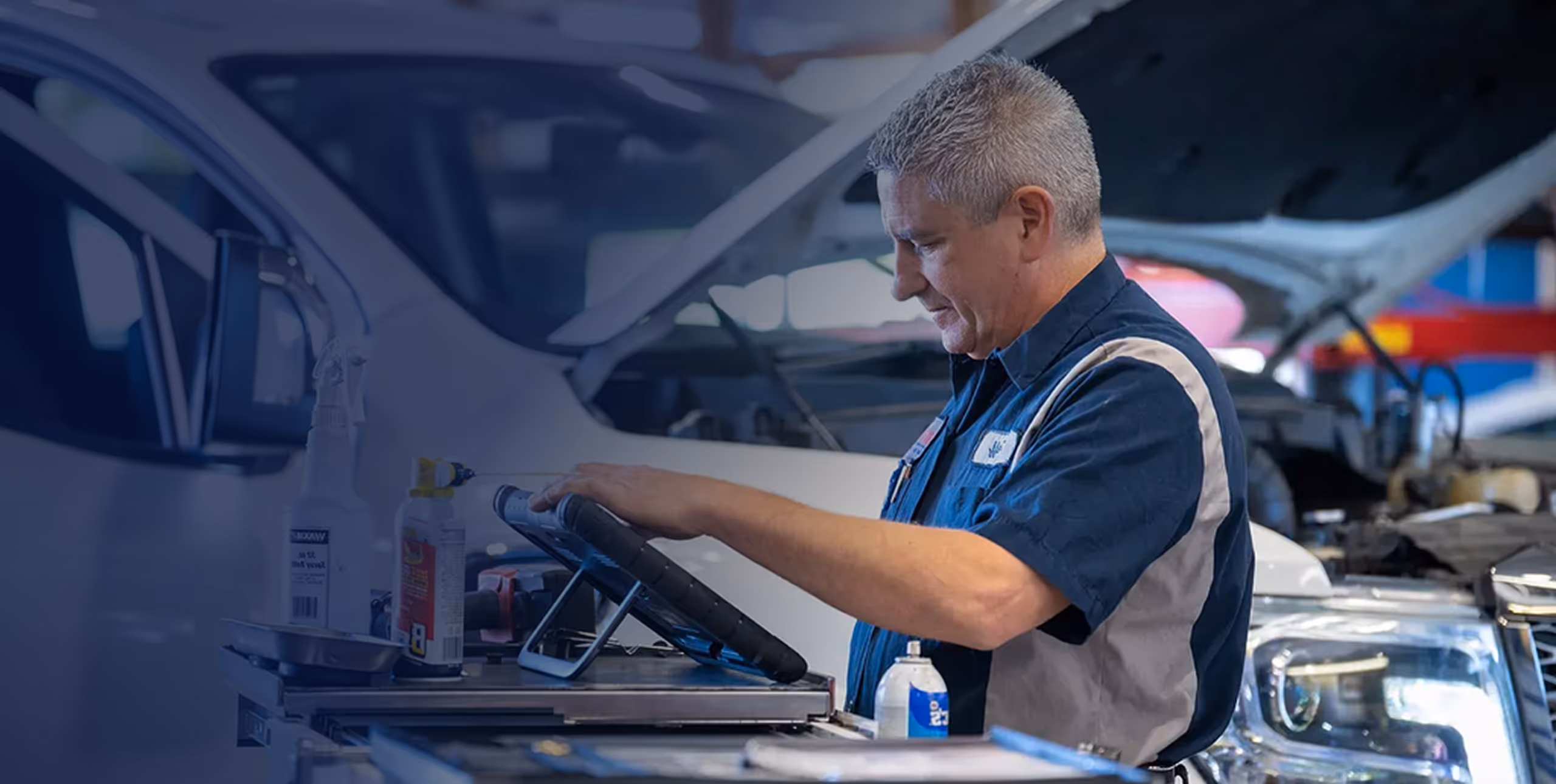 Mechanic in blue uniform using diagnostic tablet by a white car with open hood in garage.
