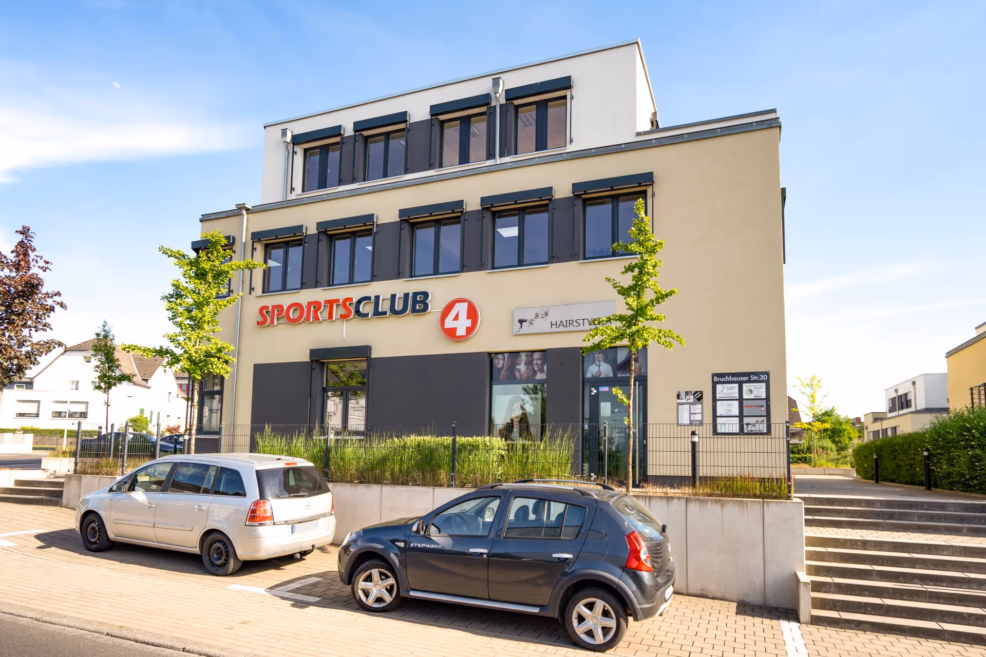 Neubau Leverkusen: Modernes, zweigeschossiges Gebäude mit der Aufschrift SPORTSCLUB 4 und zwei davor geparkten Autos unter einem klaren Himmel.