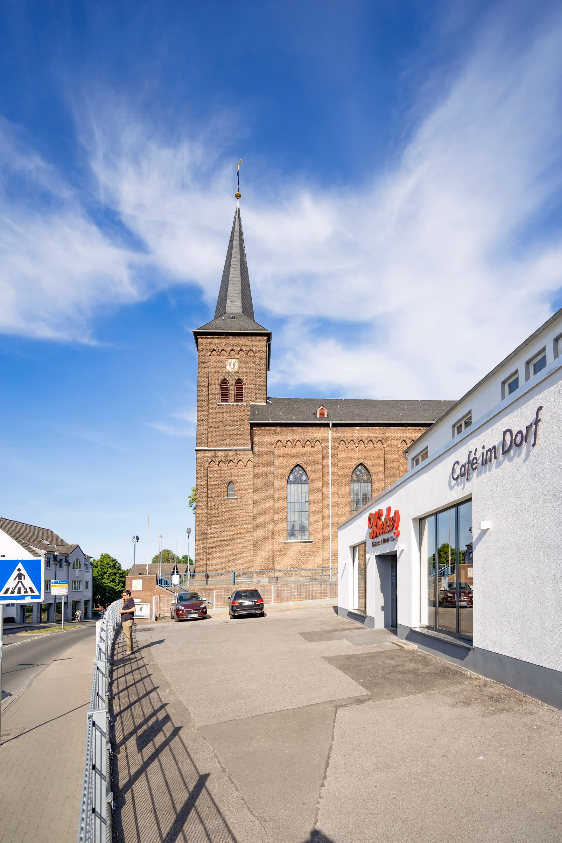 Neubau Leverkusen: Backsteinkirche mit einem hohen spitzen Kirchturm gegen einen blauen Himmel und einem weißem Gebäude mit der Aufschrift Café im Dorf auf der rechten Seite.