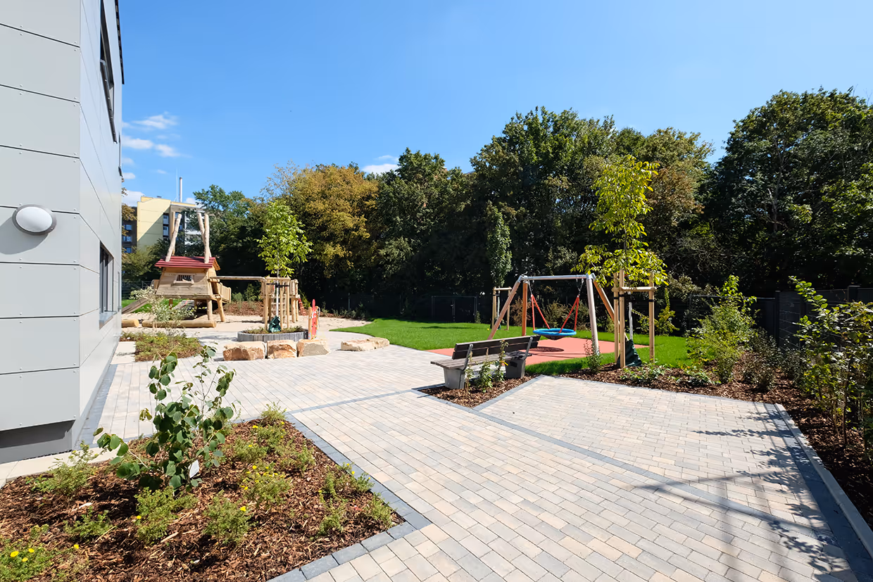 Moderner Außenspielplatz im Neubauprojekt Leverkusen Steinbüchel mit Spielgeräten aus Holz, einer Sitzbank und gepflasterten Gehwegen, umgeben von Grünanlagen unter einem klaren blauen Himmel.