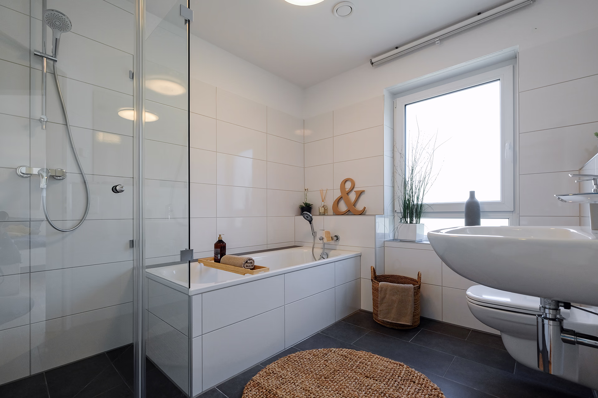 Helles, modernes Badezimmer im Neubauprojekt Leverkusen Steinbüchel mit weißen Fliesen, einer Badewanne mit Holzablage, einem Wand-WC, einem Waschbecken mit Spiegel und einer Glasdusche.