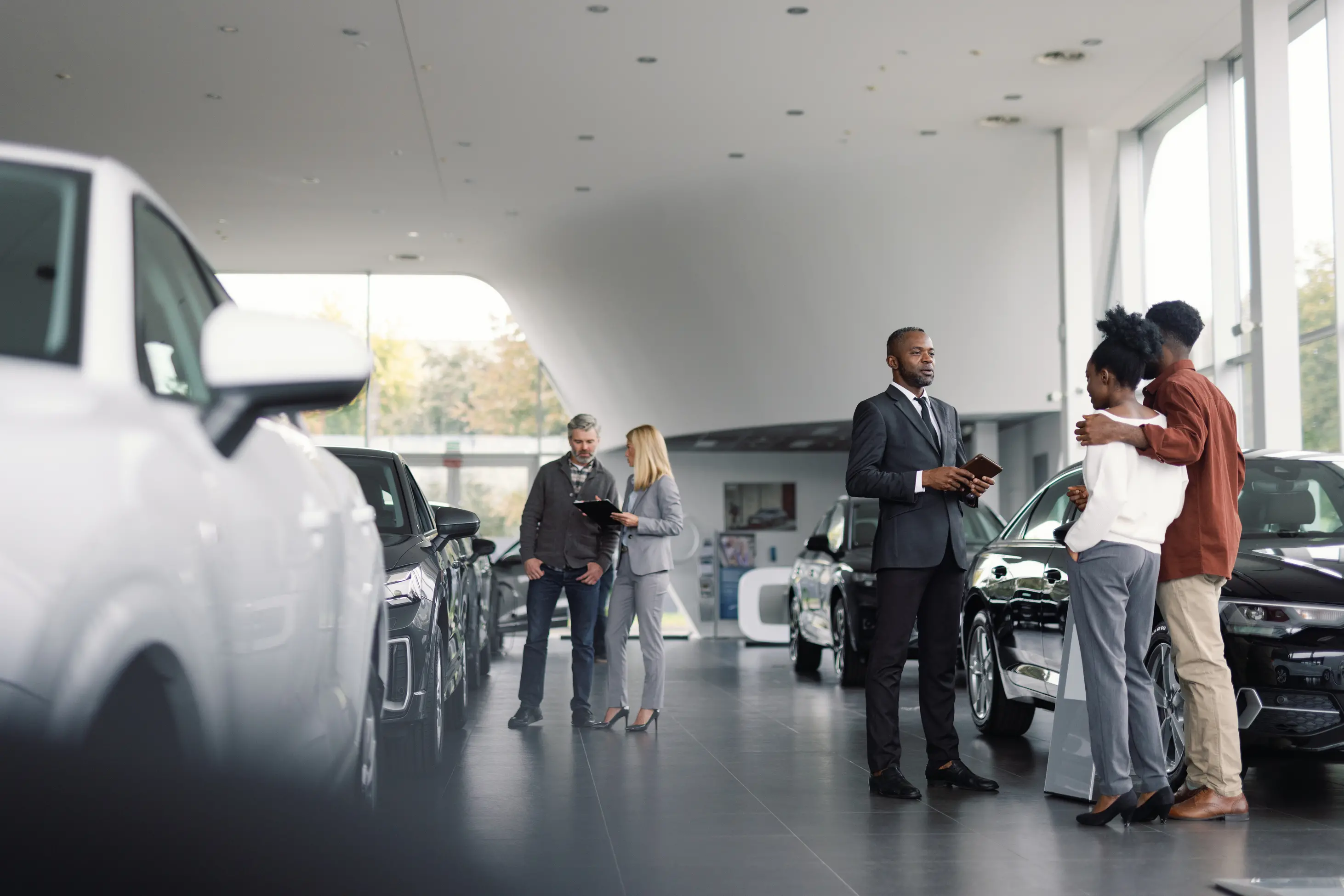 Car showroom with a couple talking to a salesperson and another couple discussing car details near vehicles.