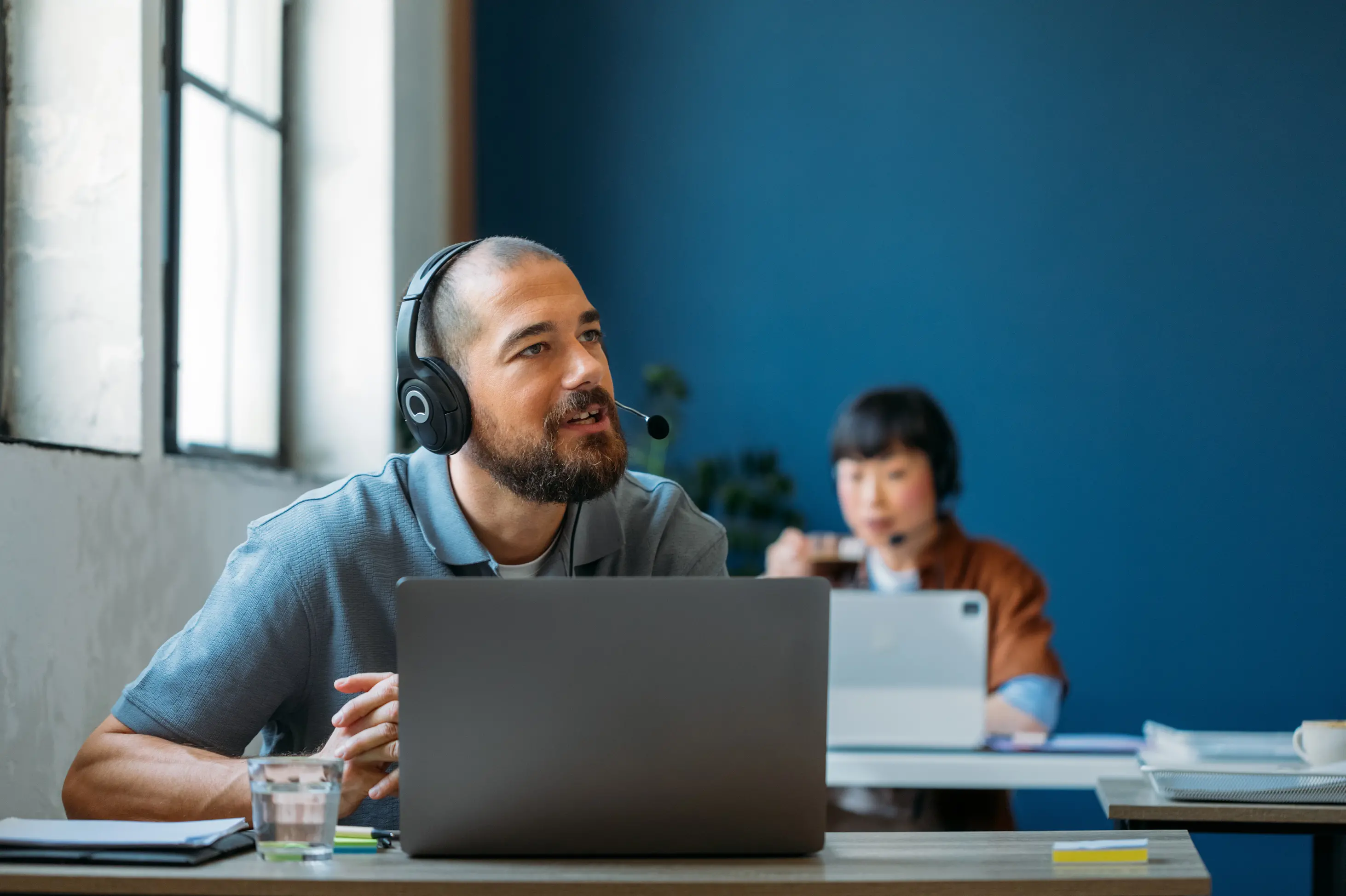 Man with headset sitting at a desk using a laptop, with a woman in the background also wearing a headset and working on a tablet.