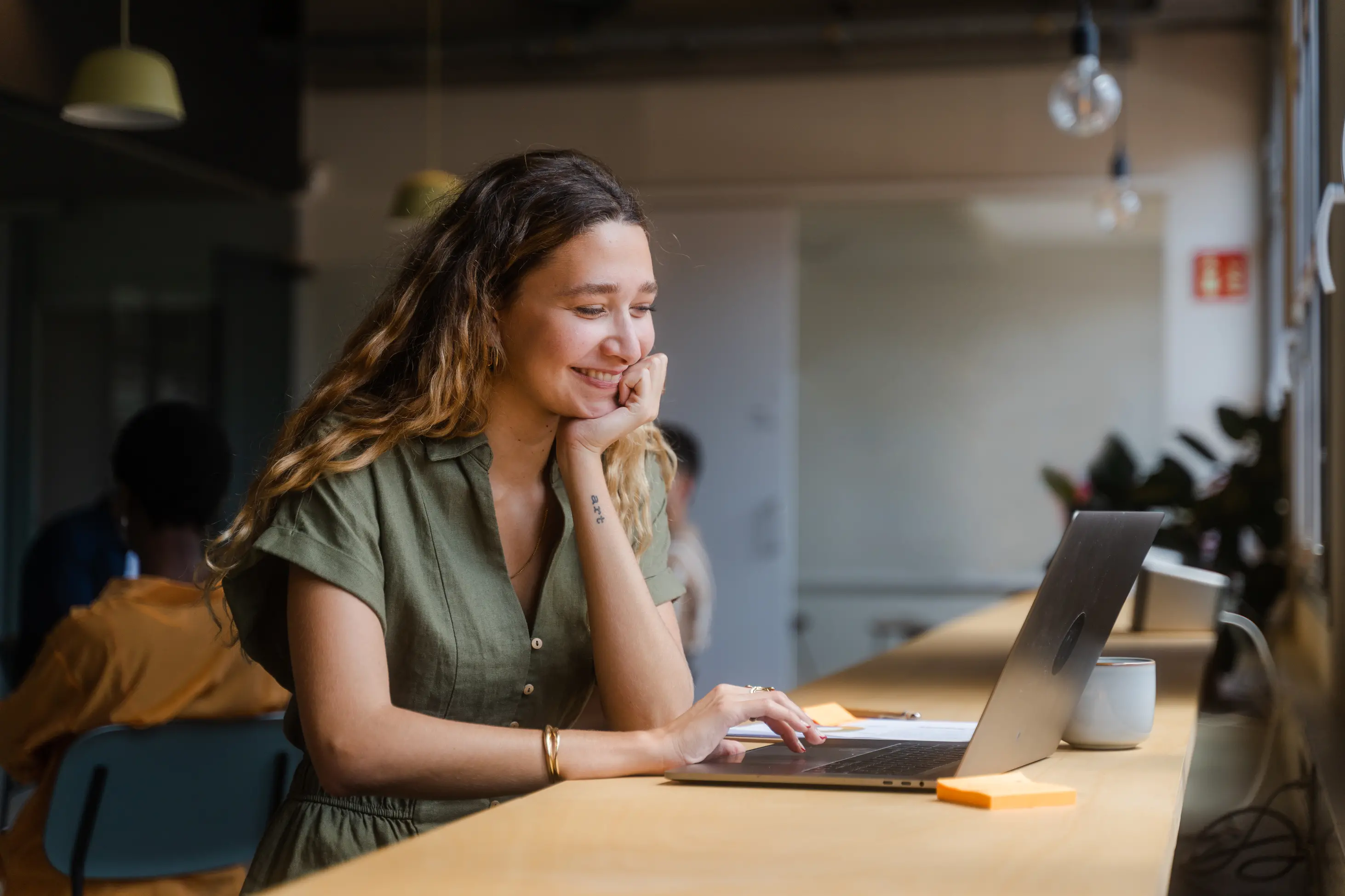 Young woman with long hair smiling while using a laptop at a wooden counter in a modern workspace.