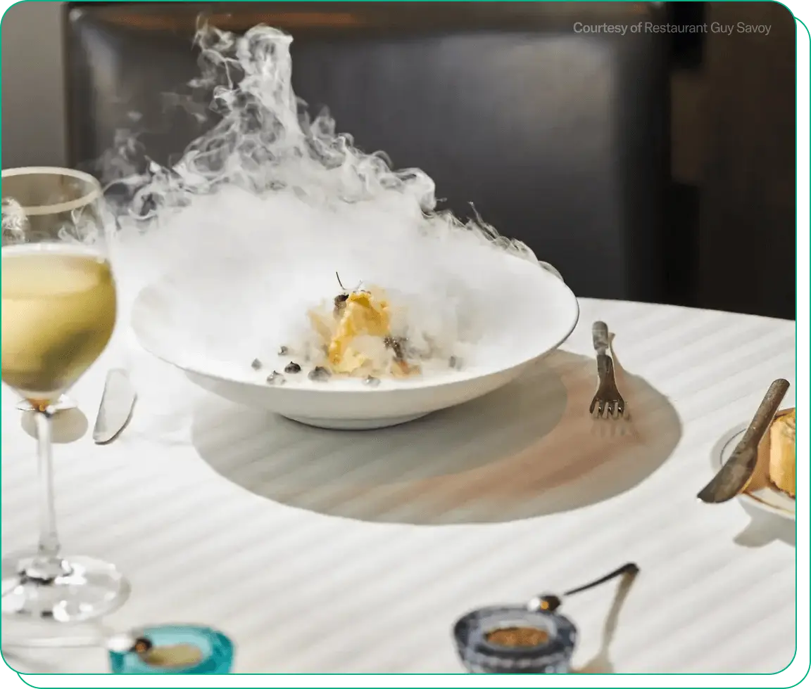 White bowl of food with smoke billowing off behind a glass of white wine