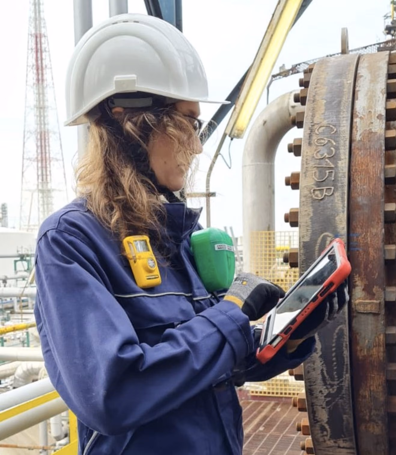 Worker in safety helmet and gloves using tablet near industrial piping and machinery.