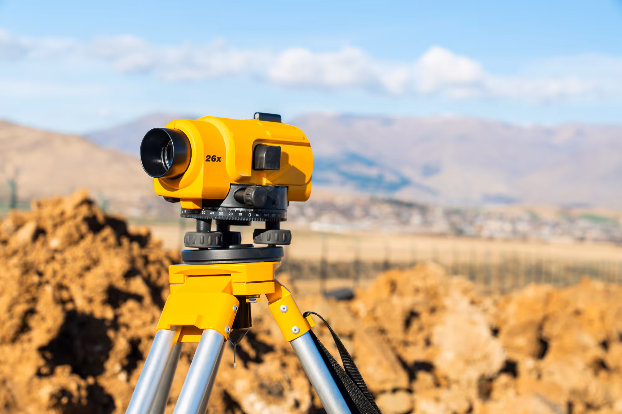 Yellow surveying theodolite with 26x zoom mounted on tripod outdoors near dirt piles.